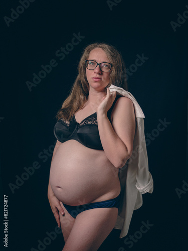 A pregnant woman standing on a dark background. She is wearing a black lace bra and blue panties, with a white shirt slung over one shoulder. Pregnancy Beauty.