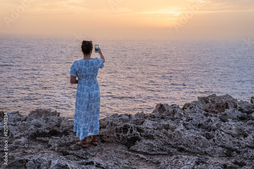 Rear view of a woman in her 30s wearing a white and blue maxi dress on a rocky shore, taking a picture with her phone of the sunset over the sea, in Menorca island, Spain