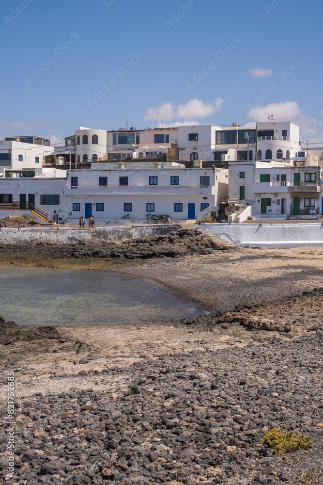 Fototapeta premium Bay and typical houses of the fishing village of Caleta de Caballo. White houses. Rock coast in the foreground. Turquoise water. Calm sea. Caleta de Caballo, Lanzarote, Canary Islands, Spain.