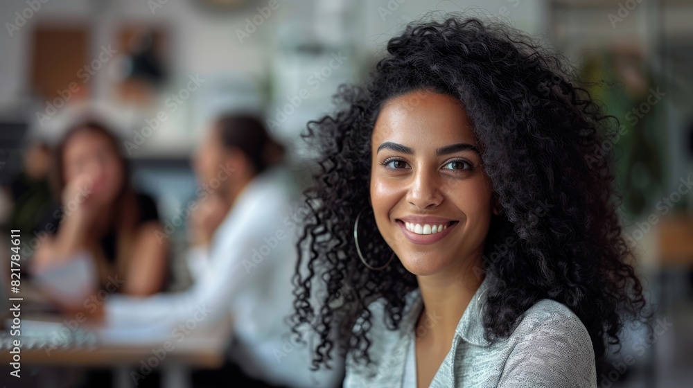 Stylish Middle Eastern Manager Sitting at Her Desk in Creative Office. Young Stylish Female with Curly Hair Looking at Camera with Big Smile. Colleagues working in the background. Stylish Middle Eastern Manager Sitting at Her Desk in Creative Office. Young Stylish Female with Curly Hair Looking at Camera with Big Smile. Colleagues working in the background.