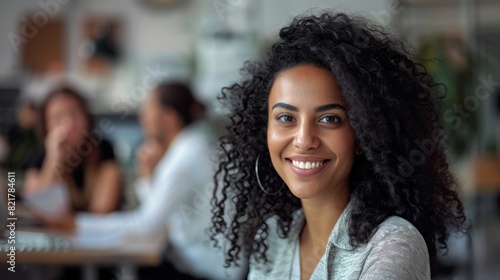 Stylish Middle Eastern Manager Sitting at Her Desk in Creative Office. Young Stylish Female with Curly Hair Looking at Camera with Big Smile. Colleagues working in the background.