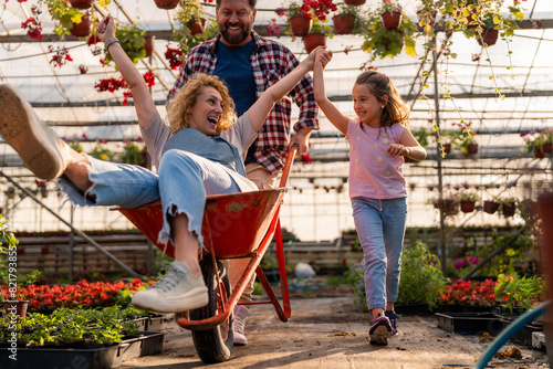 Fotografi Joyful family fun in the greenhouse, with the mother riding in a wheelbarrow pushed by her husband, while their daughter runs alongside