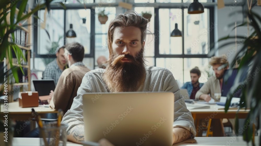 A handsome, long-haired, bearded manager sits at a desk at a marketing company, using a laptop computer. He is surrounded by colleagues. A handsome, long-haired, bearded manager sits at a desk at a marketing company, using a laptop computer. He is surrounded by colleagues.