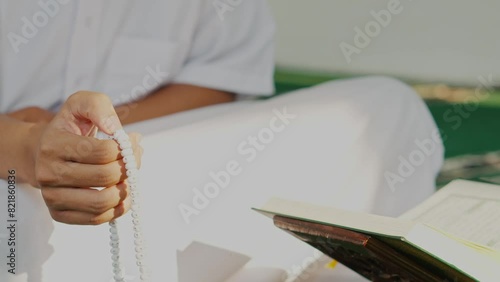 Front View Of Muslim Man Hand Holding Rosary Or Prayer Beads And Sitting Crossed Legs In Front Of Holy Quran In Mosque