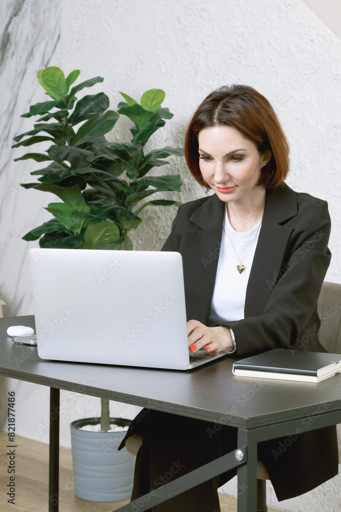 Portrait of business woman in a black business suit at desk in your office. Business portrait. Business woman is sitting in front of laptop screen Portrait of business woman in a black business suit at desk in your office. Business portrait. Business woman is sitting in front of laptop screen