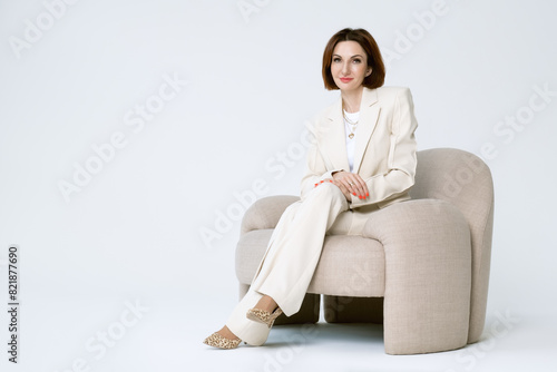 Attractive middle-aged business woman in beige pantsuit is sitting on a upholstered chair on a white background. Portrait of a business lady.