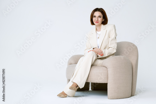 Fotografie Attractive middle-aged business woman in beige pantsuit is sitting on a upholstered chair on a white background