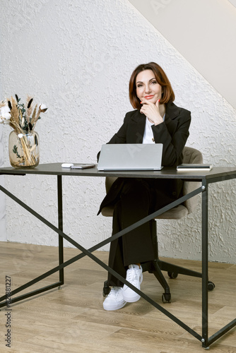 Portrait of business woman in a black business suit at desk in your office. Business portrait. Business woman is sitting in front of laptop screen