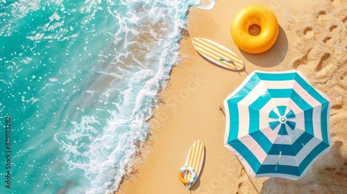 Aerial view of a sunny beach with turquoise waves, an umbrella, surfboards, and an inflatable ring on the sandy shore.