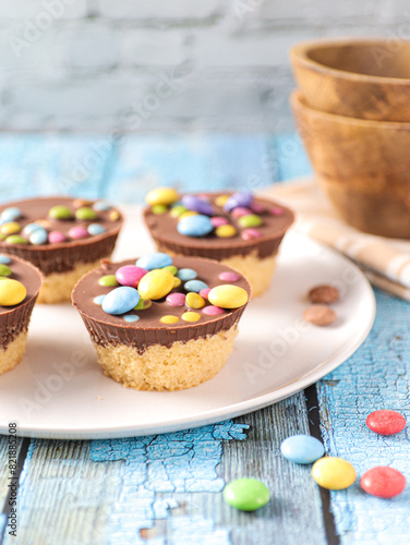 A white plate with four chocolate cupcakes topped with brown icing and colorful candies. Various colorful candies are scattered on the blue wooden surface around the plate.