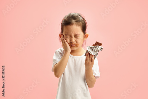 Consequences of excessive sugar consumption for children. Unhappy child touching herself cheek by dental pain standing on pink isolated background. Kid suffers from caries. photo