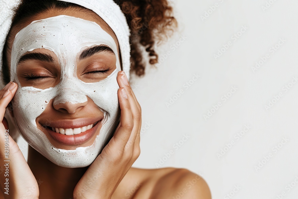 Young woman applying cream facial mask to her face gently. White bath towel on a head. Close-up. Girl is caring herself, touching cheek. Light background. Spa, body care, skincare, beauty aesthetic.