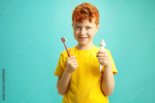 Handsome Boy holding toothpaste tube and toothbrush standing against blue isolated wall. Seven year old child using oral hygiene stuff.