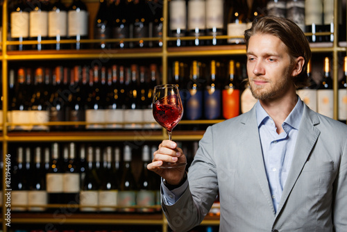 Glass of red wine on cellar background, male sommelier appreciating drink against wooden shelf with bottles.