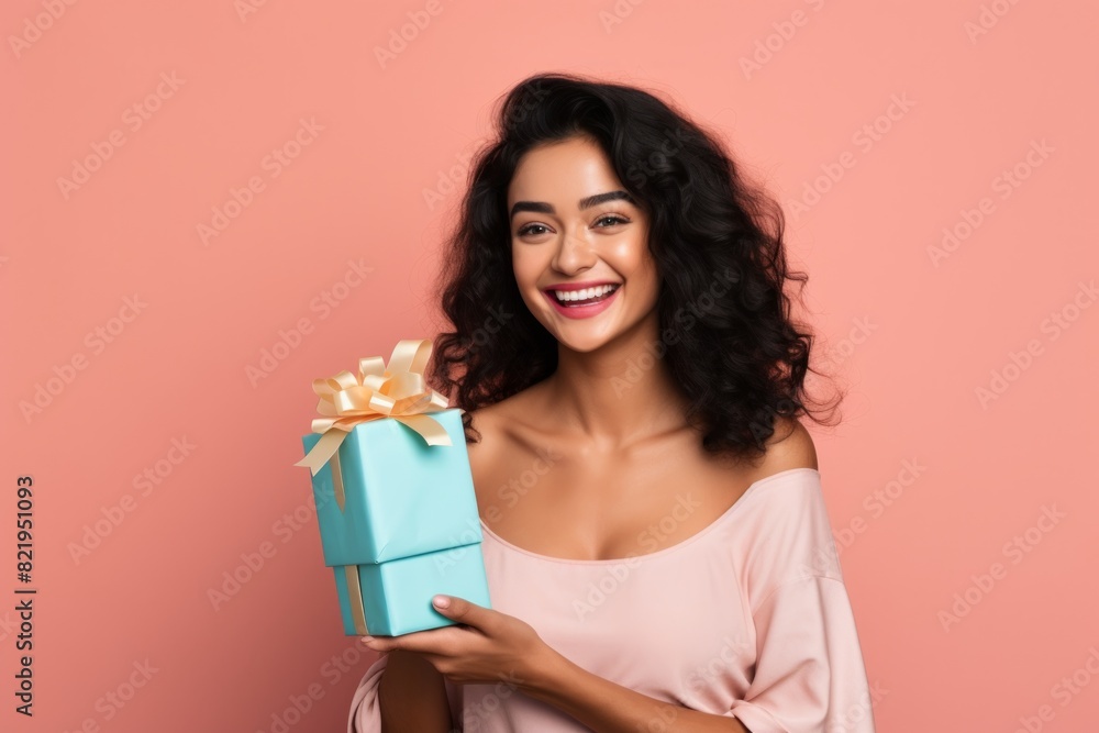 Portrait of a happy indian woman in her 20s holding a gift while standing against solid pastel color wall Portrait of a happy indian woman in her 20s holding a gift while standing against solid pastel color wall