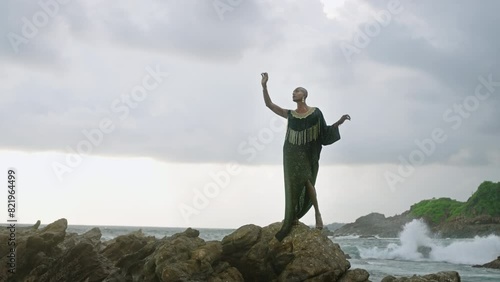 Queer black person gracefully poses in posh dress, handmade jewelry stands on scenic sea cliff top above dramatic cloudy sky, ocean waves. Lgbtq biethnic gay model in brass accessories. photo