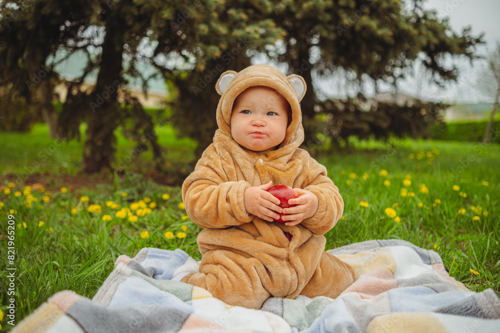Cute baby girl in plush suit with red apple in hands sitting on blanket in green grass. Cute baby girl in plush suit with red apple in hands sitting on blanket in green grass.