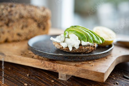 Fresh avocado slices on grain bread with cream cheese and lemon. Vegetarian food on wooden cutting board. Close-up in front of bright window. Short depth of field.