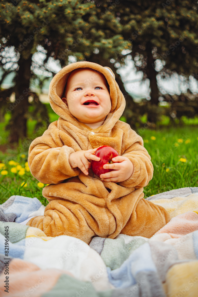 Cute baby girl in plush suit with red apple in hands sitting on blanket in green grass. Cute baby girl in plush suit with red apple in hands sitting on blanket in green grass.