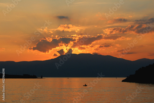 Lone old fisherman fishing for his family in a small boat at local bay on calm sea in front of tall mountain at sunset with cloudy sky in background