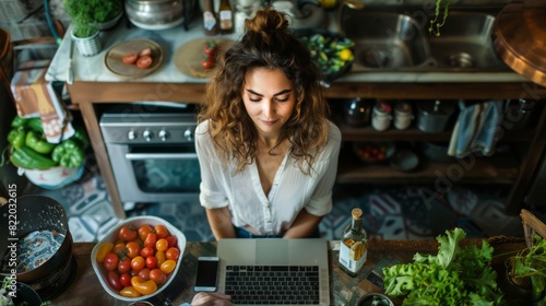 Young woman working on a laptop in a cozy kitchen surrounded by fresh vegetables and fruits, practicing healthy eating and remote work.