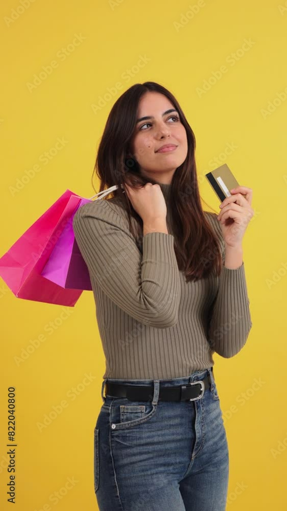 Vertical portrait of a young caucasian customer woman holding shopping bags and a credit card, contemplating whether to buy clothes from an online store. She is against a yellow background purchasing Vertical portrait of a young caucasian customer woman holding shopping bags and a credit card, contemplating whether to buy clothes from an online store. She is against a yellow background purchasing