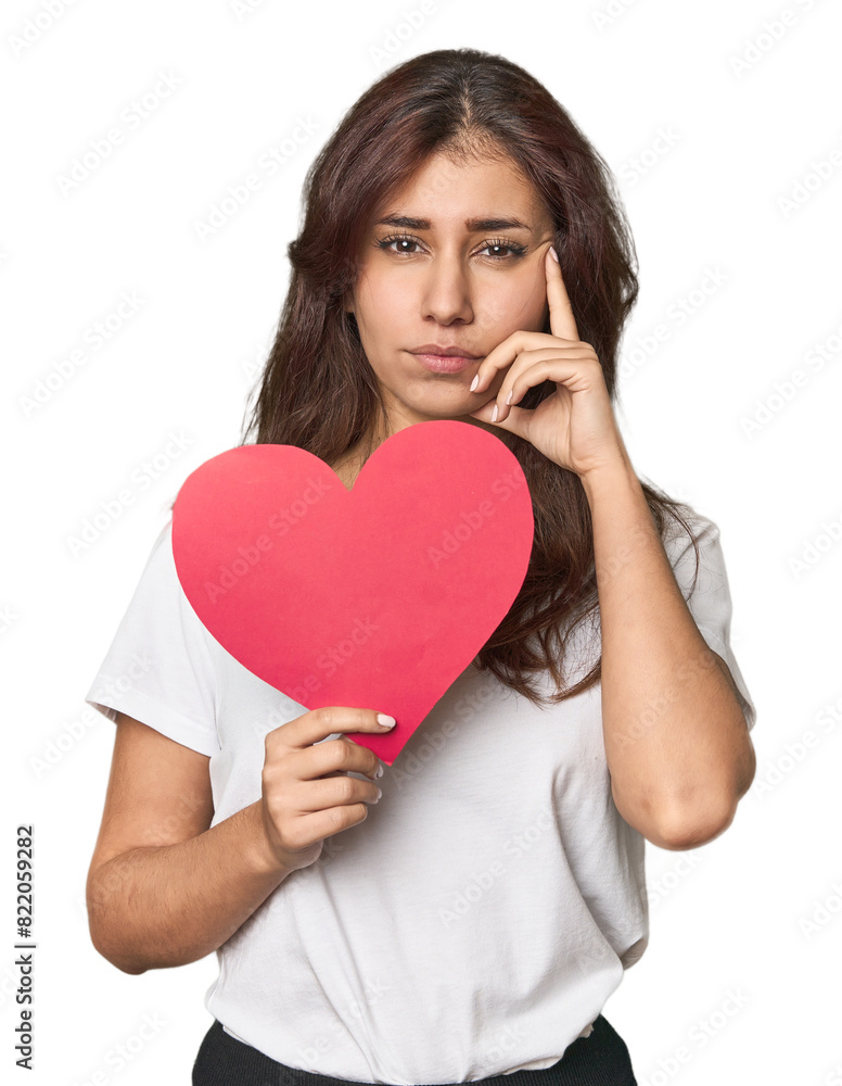 Middle Eastern woman holding paper heart pointing temple with finger, thinking, focused on a task. Middle Eastern woman holding paper heart pointing temple with finger, thinking, focused on a task.