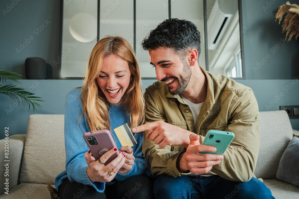 © Jose Calsina - Young female holding a credit card, shopping online and her husband pointing her smart phone. Adult couple searching a sale on a cellphone social media app sitting on a couch at home living room © Jose Calsina - Young female holding a credit card, shopping online and her husband pointing her smart phone. Adult couple searching a sale on a cellphone social media app sitting on a couch at home living room