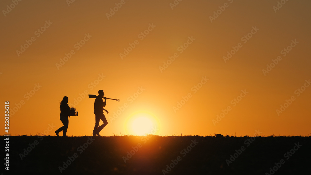 Two farmers with work equipment walk through a plowed field at sunset