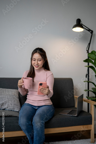 A woman is sitting on a couch, holding a pink mug and a cell phone. She is smiling and she is enjoying her time