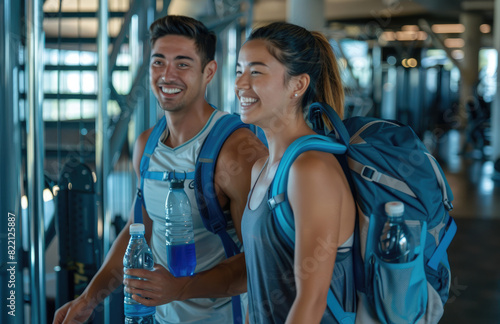 A man and woman walking out of the gym, smiling at each other with sportswear on their shoulders and fitness equipment in the background