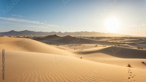 This is an image of a desert with a sunrise or sunset. There are sand dunes in the foreground and mountains in the background. The sky is clear with a bright sun.  