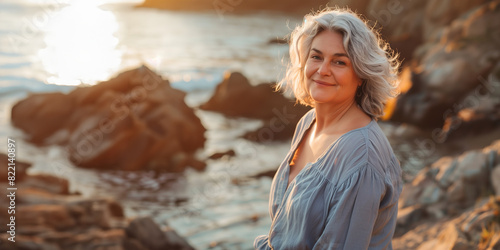 60-year-old woman with silver hair on a beach, dressed in sustainable linen attire against rocky seascapes at sunset. Concept of affluent retirement, pension travel, travel insurance
