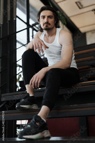 A confident man in a white tank top and black pants sits on stairs in a modern building  looking directly at the camera.