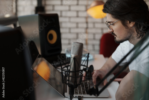Focused man working on a laptop in a home recording studio with a microphone setup, surrounded by audio equipment. photo