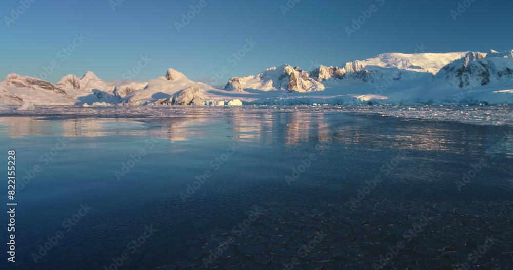 Explore icy wonders of Antarctica winter landscape. Snow covered mountain range in sunset light, cold ice ocean water. Beauty of wild untouched nature. Arctic scene. Low angle drone flight