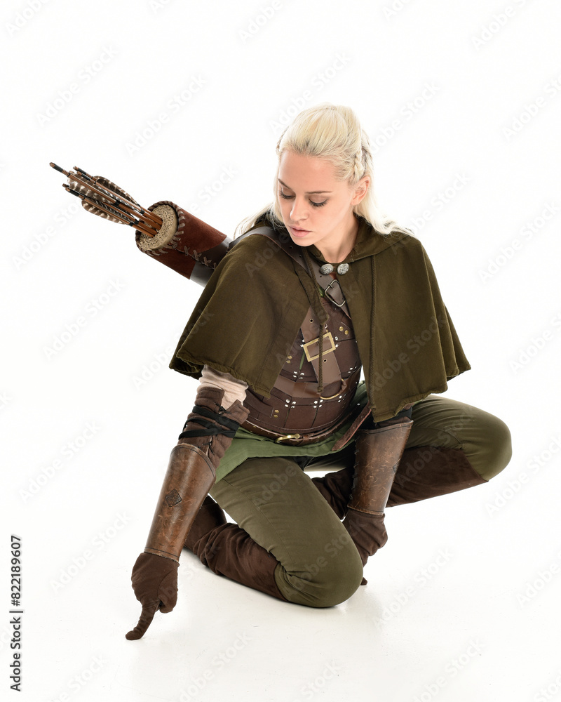 full length portrait of blonde female model wearing green fantasy elf warrior halloween costume with leather armour. sitting pose kneeling. isolated on white studio background. full length portrait of blonde female model wearing green fantasy elf warrior halloween costume with leather armour. sitting pose kneeling. isolated on white studio background.