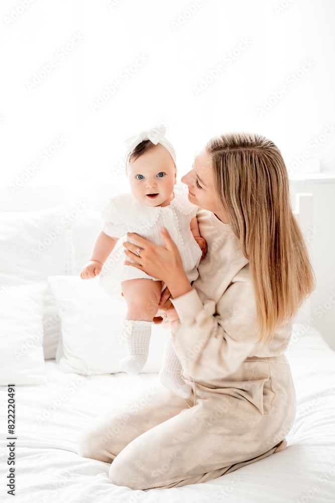 a mother with a newborn baby in her arms at home on a white background of a bed or window, a space for text, a young mother gently kisses and hugs her baby, maternal love a mother with a newborn baby in her arms at home on a white background of a bed or window, a space for text, a young mother gently kisses and hugs her baby, maternal love