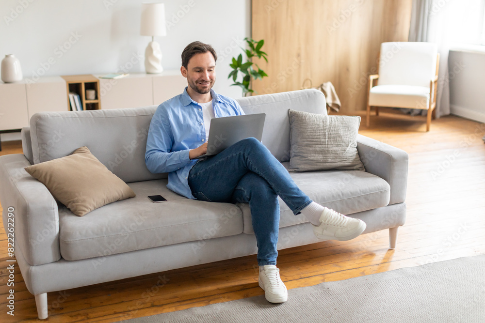 A man is sitting comfortably on a couch, focused on his laptop screen. He appears engaged in typing or browsing online content. The room is well-lit, with various items around him. A man is sitting comfortably on a couch, focused on his laptop screen. He appears engaged in typing or browsing online content. The room is well-lit, with various items around him.
