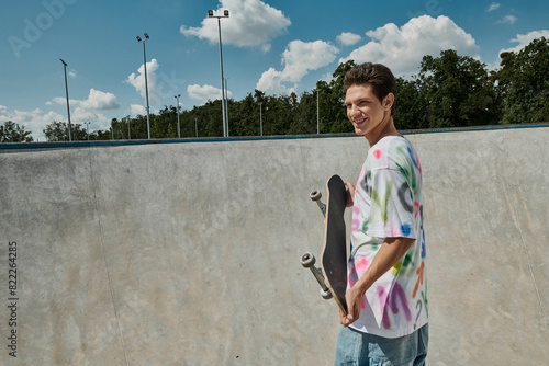 A young man confidently holds his skateboard in a vibrant skate park on a sunny summer day.