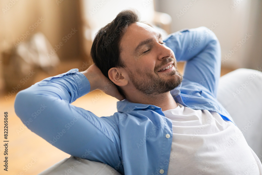 A man is seated on a couch, leaning back with his hands on his head. He appears relaxed and contemplative as he takes a moment to unwind at home.