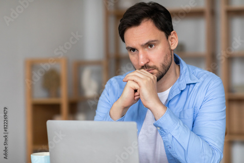 A man is seated in front of a laptop, deep in thought, with his hand resting on his chin. He appears to be contemplating or pondering something while working on his computer.