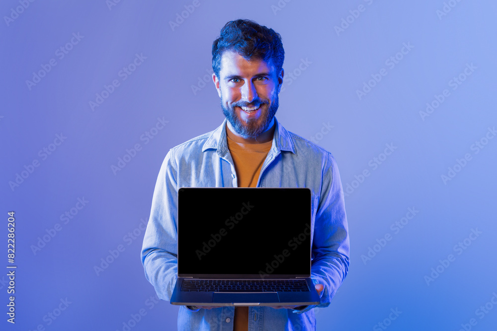 A young bearded man wearing a casual shirt smiles while presenting a laptop with a blank screen. The background is blue, creating a cool ambiance. A young bearded man wearing a casual shirt smiles while presenting a laptop with a blank screen. The background is blue, creating a cool ambiance.