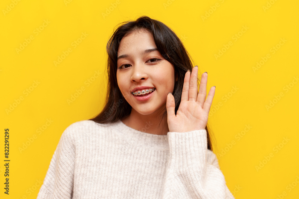 young asian girl with braces greets and shows palm of hand on yellow isolated background, korean girl shows hello and bye gesture young asian girl with braces greets and shows palm of hand on yellow isolated background, korean girl shows hello and bye gesture