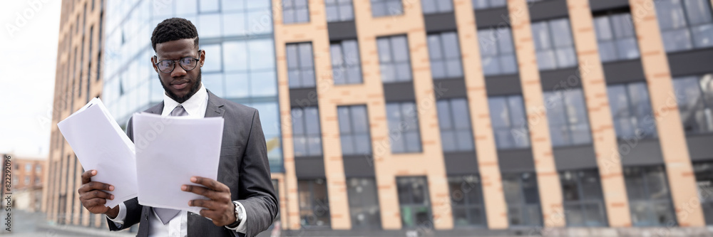 Young black man in suit checking reports or getting ready for job interview, holding documents or CV, standing outdoors against office center, copy space Young black man in suit checking reports or getting ready for job interview, holding documents or CV, standing outdoors against office center, copy space