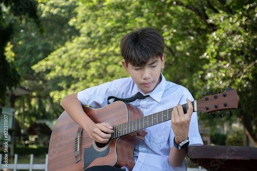 Asian preteen schoolboy in school uniform sitting and playing acoustic guitar in schoolpark, recreational activity of children concept.