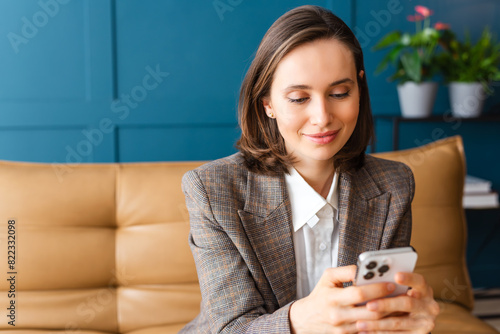 Beautiful woman with brown hair sitting on the sofa, smiling and reading a message on her mobile phone.