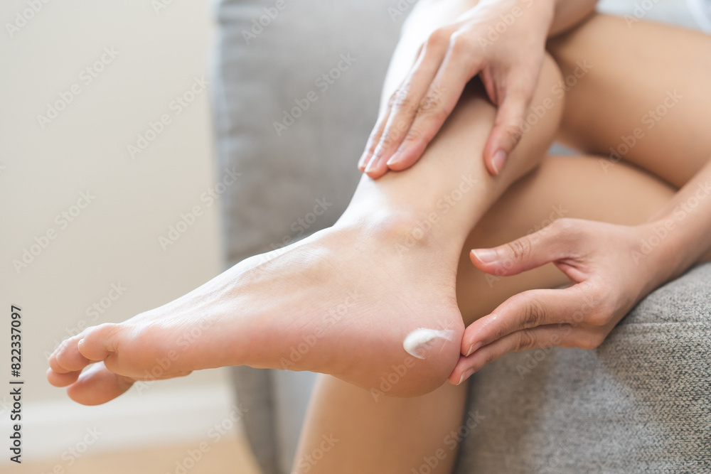 Fototapeta premium Foot care, healthcare concept, beauty asian young woman, girl hand applying moisturizing massage cream for feet dry skin on heel with lotion sitting on sofa at home, moisturizer, treatment cosmetic.