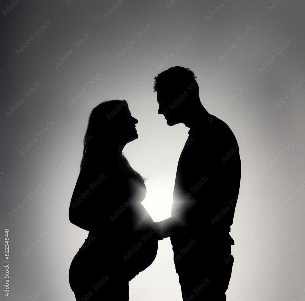 Sunlight shining behind silhouette of pregnant woman facing a man as he touches her stomach; Studio Shot Sunlight shining behind silhouette of pregnant woman facing a man as he touches her stomach; Studio Shot
