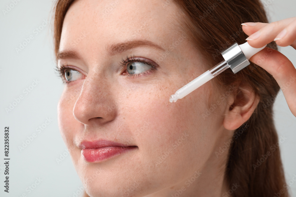 Beautiful woman with freckles applying cosmetic serum onto her face against grey background, closeup